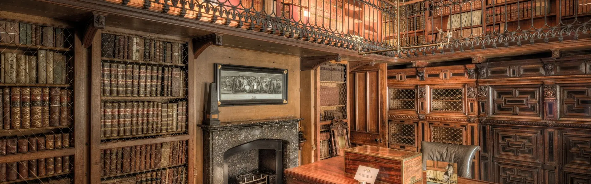 Biblioteca antigua de dos alturas, con estanterías de madera acristaladas, galería de hierro forjado, chimenea de mármol y escritorio en el centro
