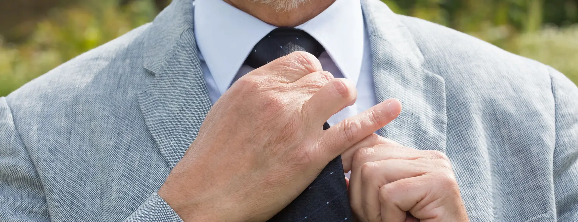 Primer plano de un caballero de barba blanca ajustándose el nudo de la corbata, con americana clara de tejido de lino
