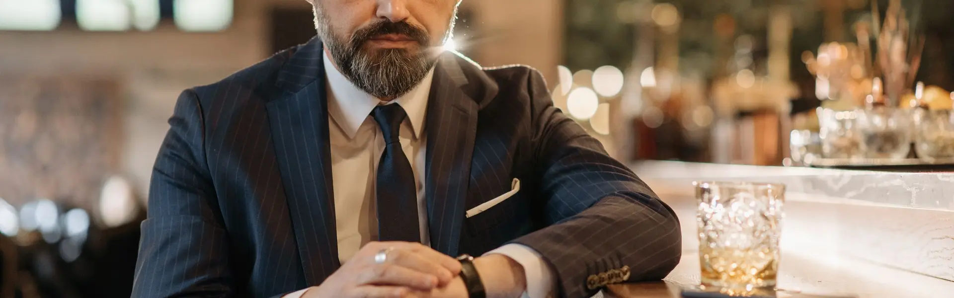 Caballero con traje azul de raya diplomática consultando su reloj en la barra de un bar