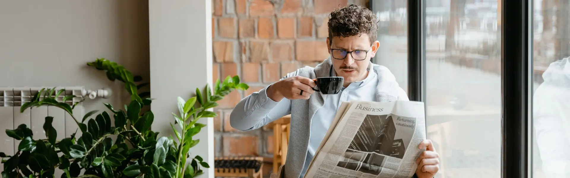 Hombre leyendo la sección de economía del periódico con una taza de café junto a un ventanal