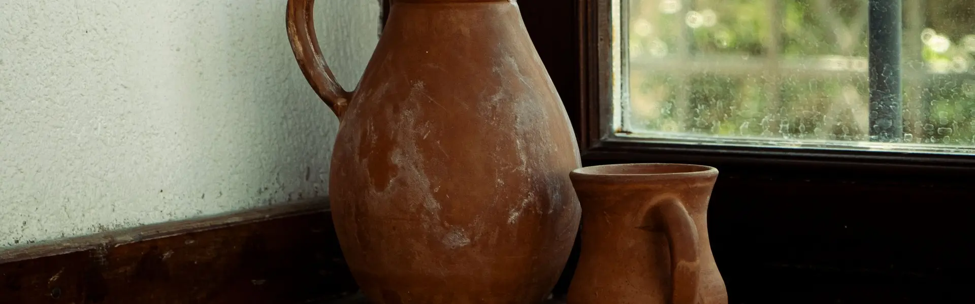 Cántaro de barro con su tapa y una taza de arcilla sobre el poyo de madera de una ventana enrejada