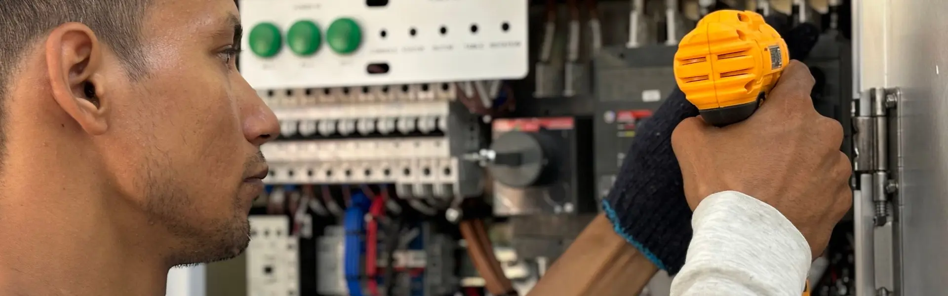 Electricista atornillando con el taladro en un cuadro eléctrico abierto, junto a la botonera de mandos