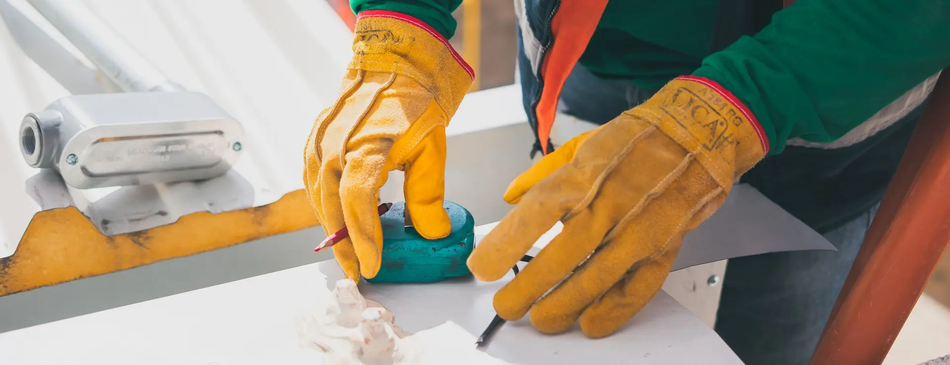 Manos con guantes de cuero amarillos marcando mediciones con lápiz sobre la chapa de un tejado a pleno sol