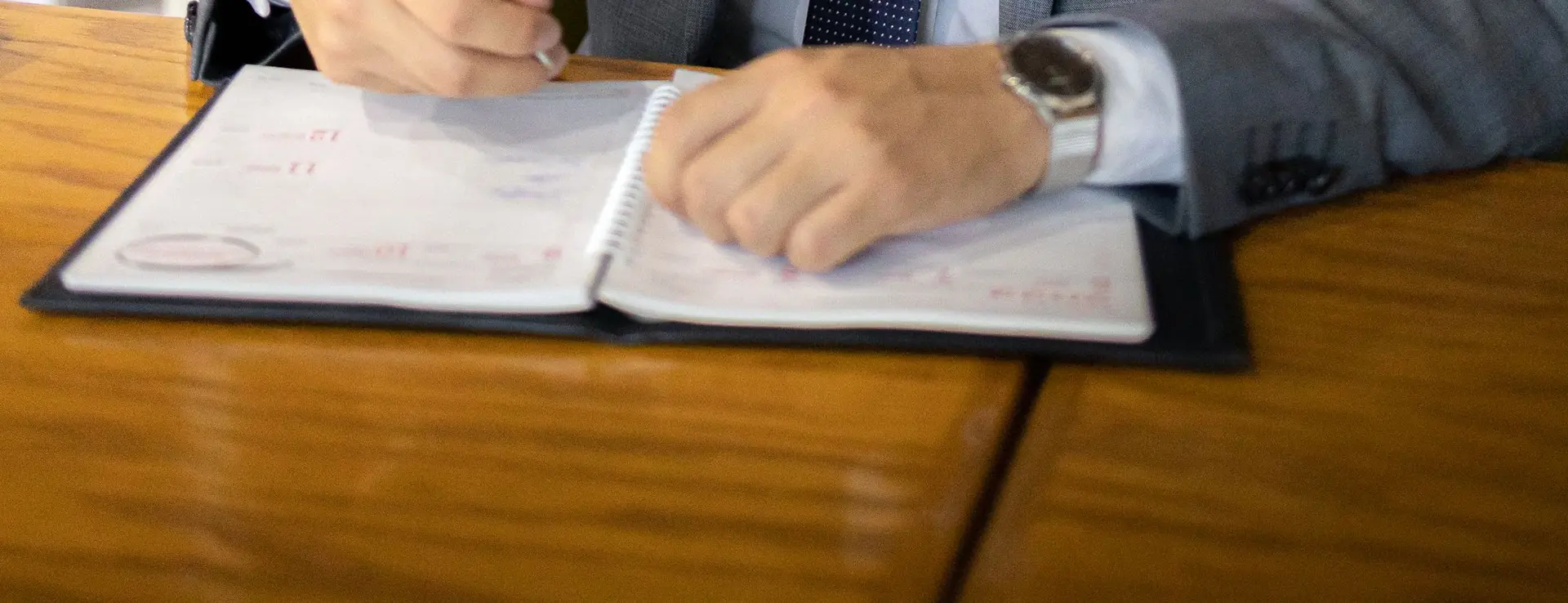 Hombre joven con traje gris y corbata, pluma en mano, sentado a su escritorio ante una estantería de libros