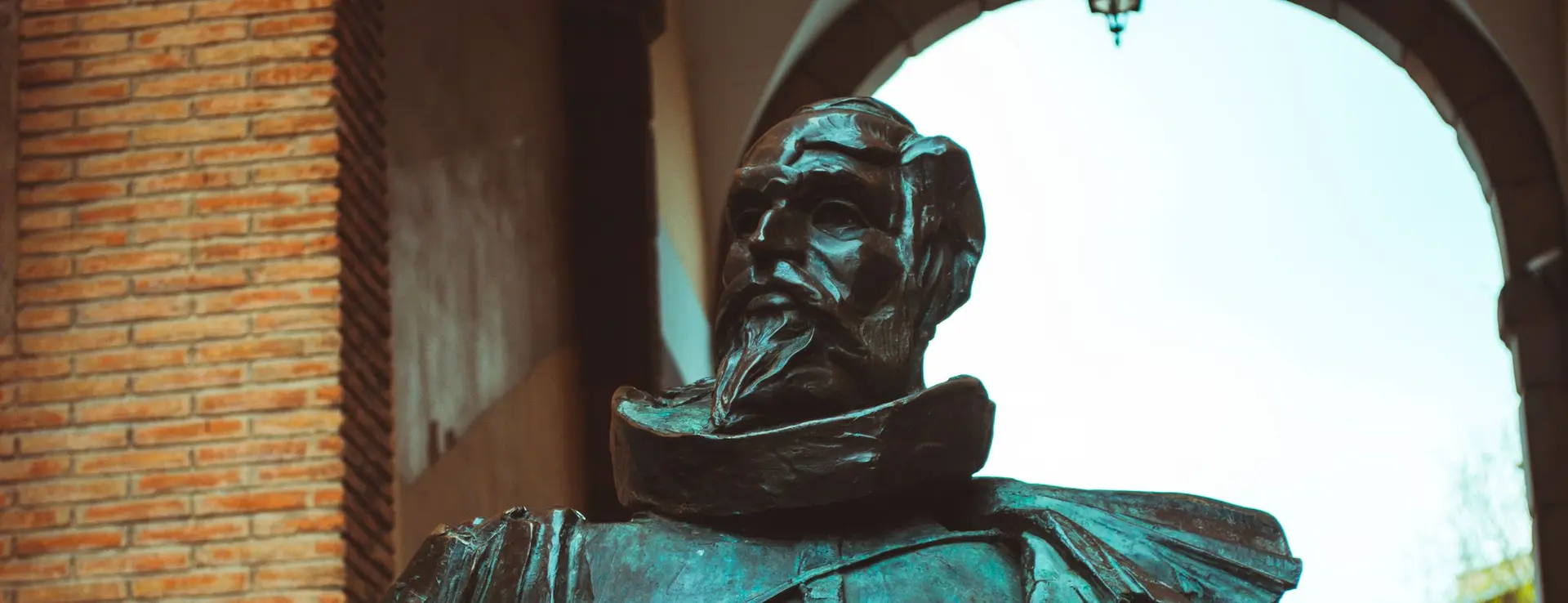 Estatua de bronce de un caballero español con gorguera y barba, sosteniendo un libro, bajo un arco de Toledo