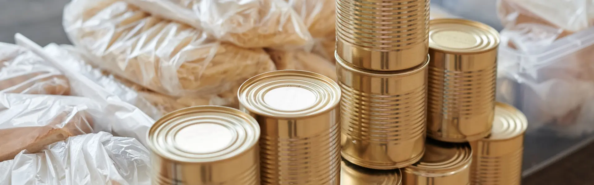Latas de conserva doradas, sin etiqueta, apiladas sobre una mesa de madera junto a barras de pan embolsadas