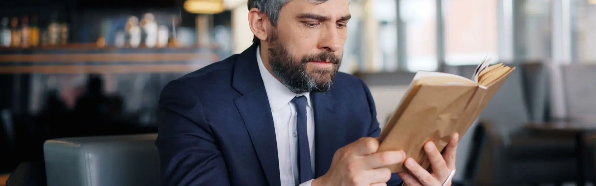 Hombre maduro de barba cana y traje azul marino leyendo un libro en una cafetería