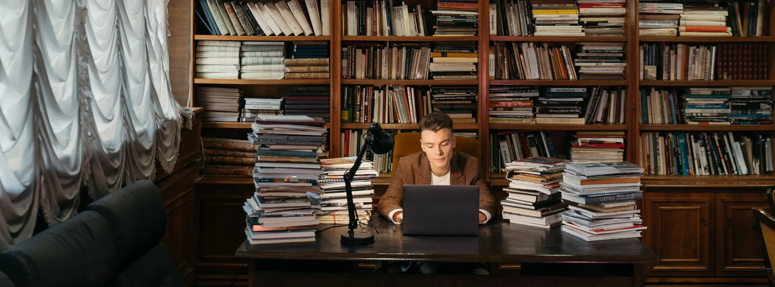 Joven estudiando con su portátil entre pilas de libros en una biblioteca clásica