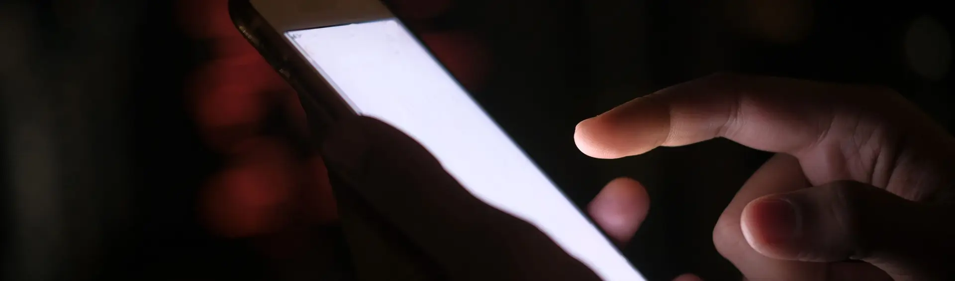 Mano pulsando la pantalla encendida de un teléfono en la oscuridad, con luces de ciudad desenfocadas al fondo