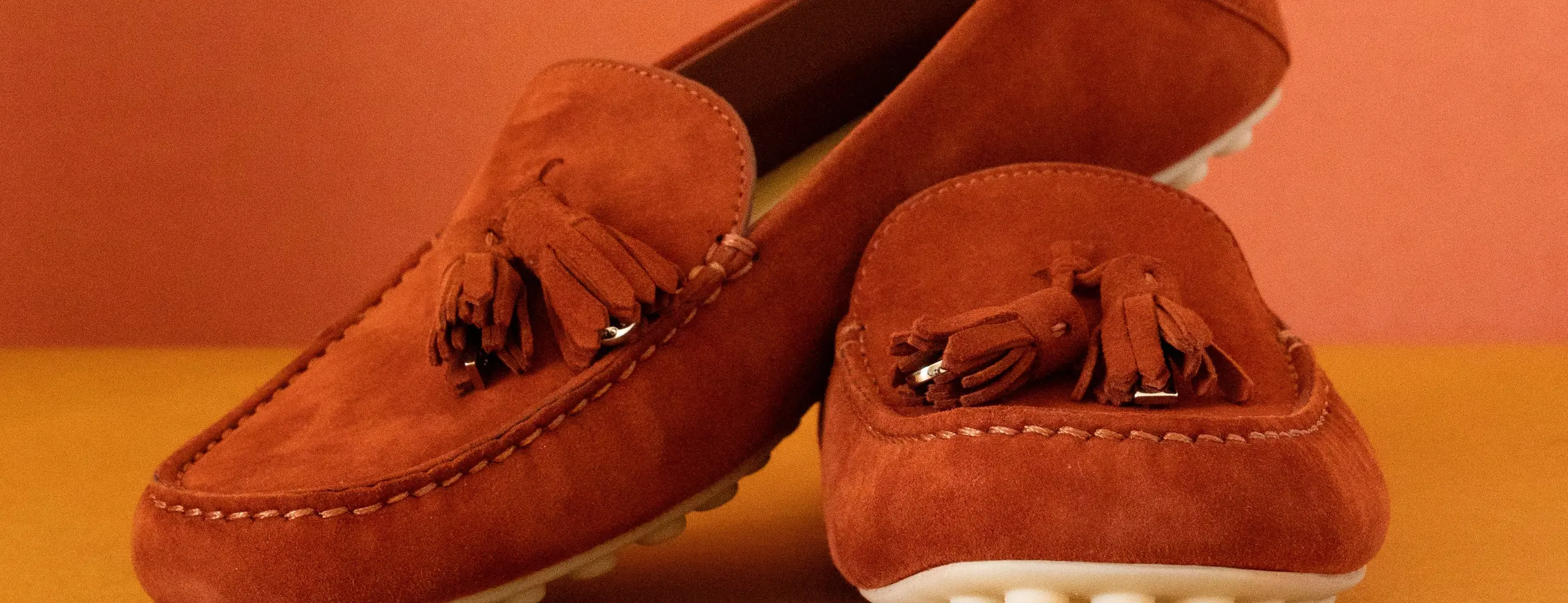 Mocasines de conducir de ante color teja con borlas, sobre fondo naranja y ocre