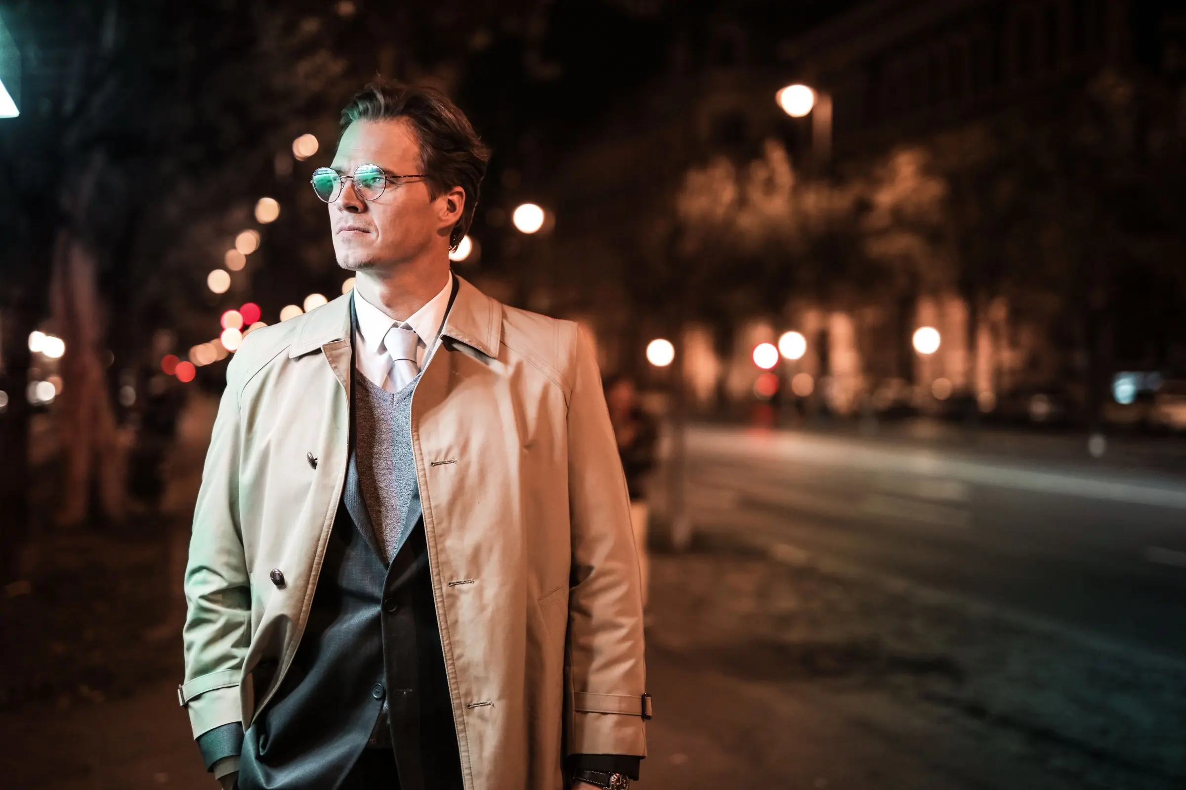 Caballero con gabardina, corbata y gafas en una avenida nocturna
