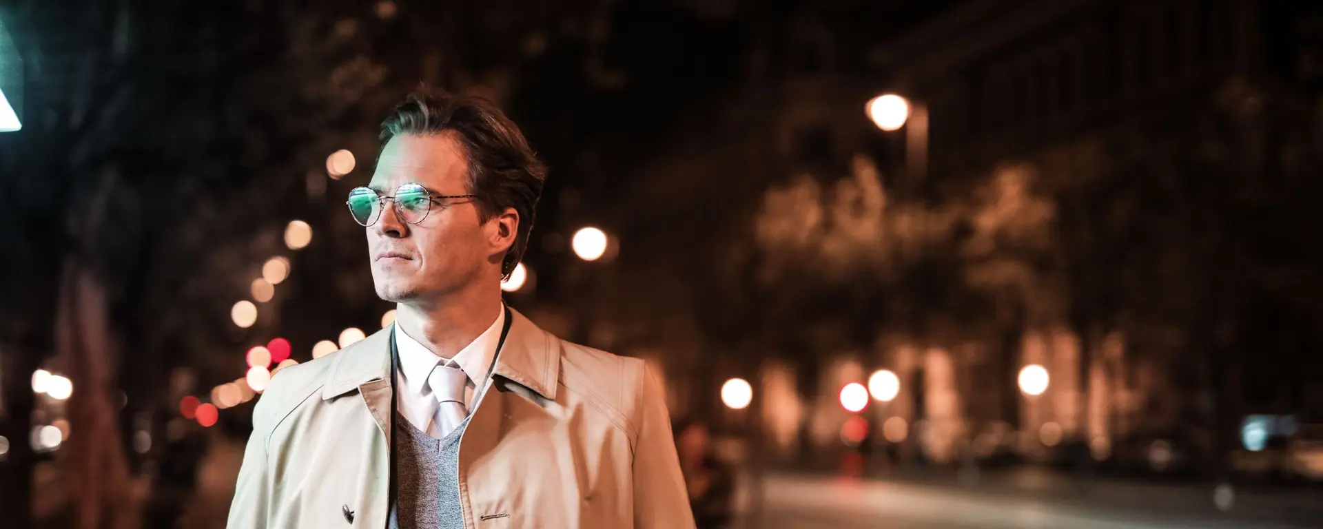 Caballero con gabardina, corbata y gafas en una avenida nocturna