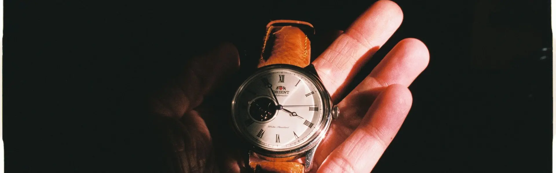 Mano sosteniendo un reloj Orient clásico de correa de piel marrón bajo una luz dramática