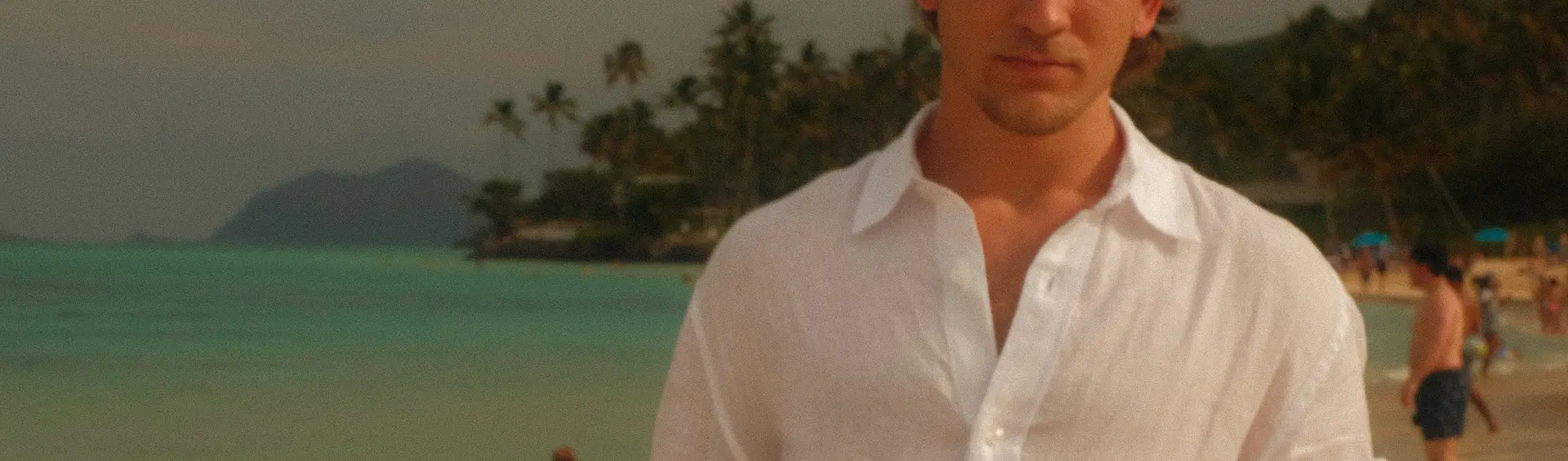 Hombre con camisa de lino blanca y pantalón de lino color arena, junto a la orilla al atardecer