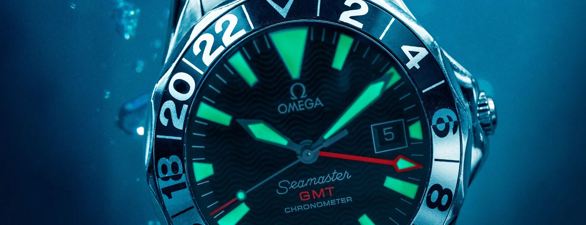 Reloj de buceo de esfera negra e índices luminiscentes, sumergido en agua azul entre burbujas
