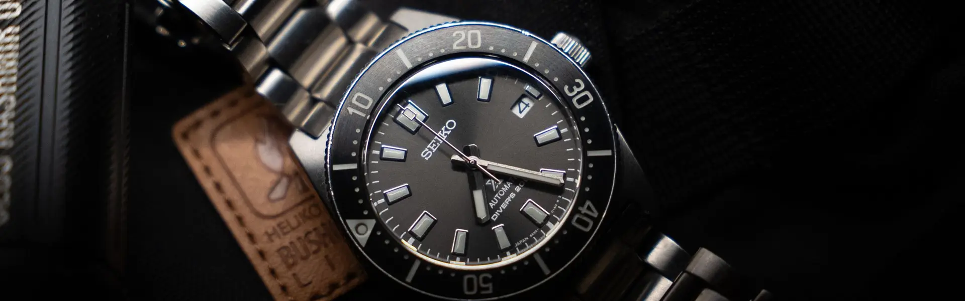 Reloj de buceo Seiko de esfera gris y bisel negro sobre brazalete de acero, junto a una linterna, sobre tela oscura