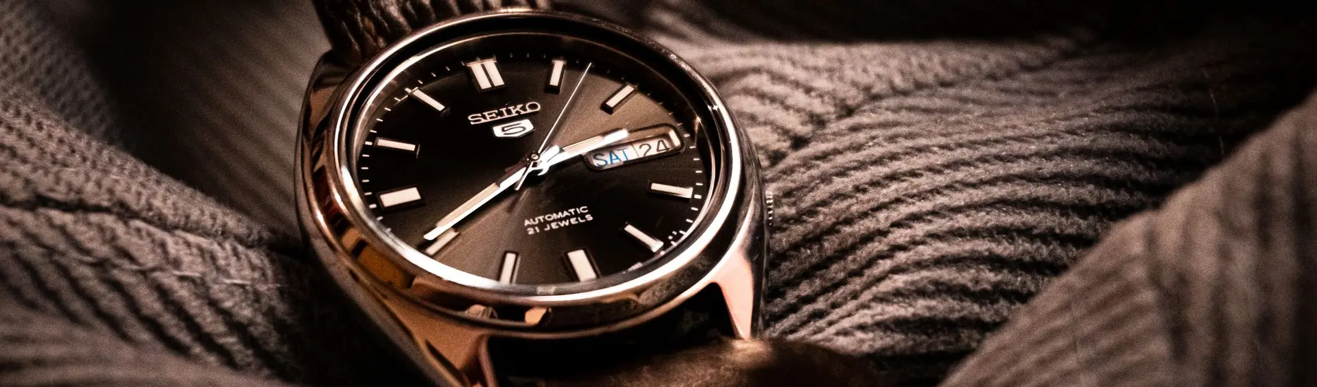 Seiko 5 clásico de esfera negra con calendario, posado sobre un jersey de punto gris