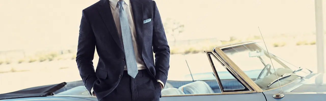 Caballero con traje de lino azul marino, camisa blanca, corbata clara y pañuelo de bolsillo, apoyado en un descapotable clásico